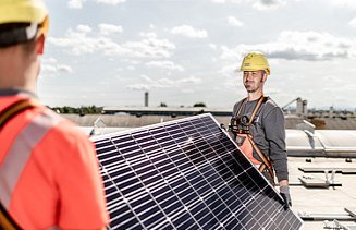 Foto von Bauarbeitern, die ein Solarpanel tragen, auf dem Solarzellen gut erkennbar sind. 